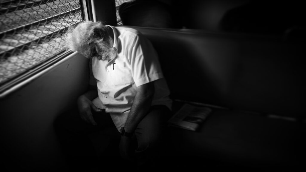 monochrome black and white photo of old man sleeping at window seat in mumbai local train man with white shirt and beard with grey hair